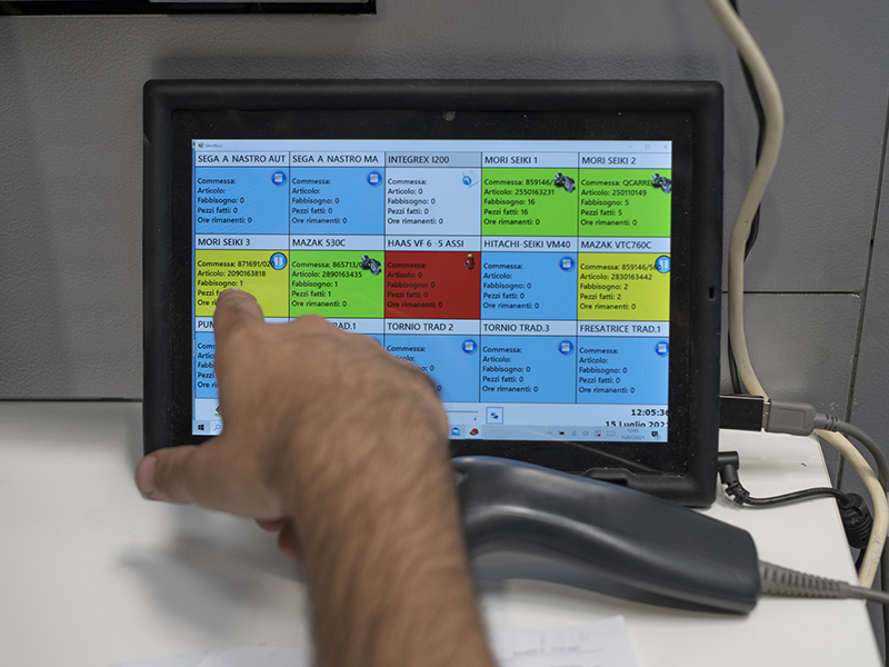 Una mano indica lo schermo di un tablet, dove sono riportate tante caselle colorate in vari colori a rappresentare le macchine in lavoro e la relativa commessa.